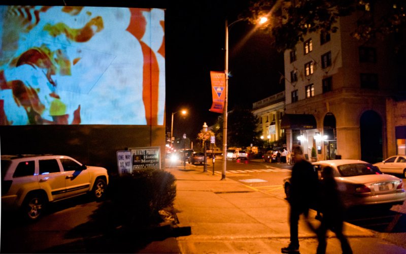 Nighttime street scene with abstract blue, orange, and white projection on a building, cars parked, and people walking.