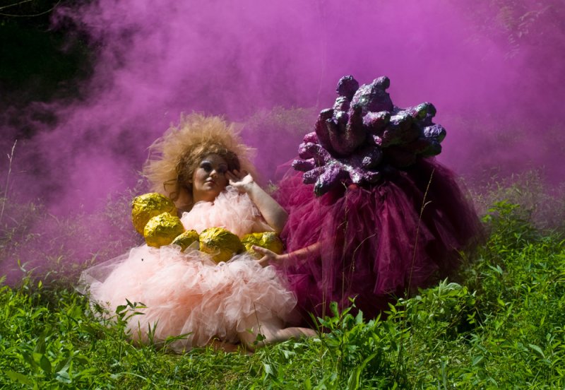 Two figures in elaborate costumes, one in a pink ruffled dress with gold objects, the other in a purple textured gown, amidst