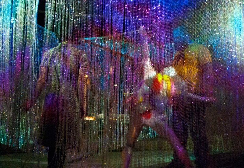 Three figures, one upside down, obscured by shimmering, vertical strands of light in purple, blue, and green hues.