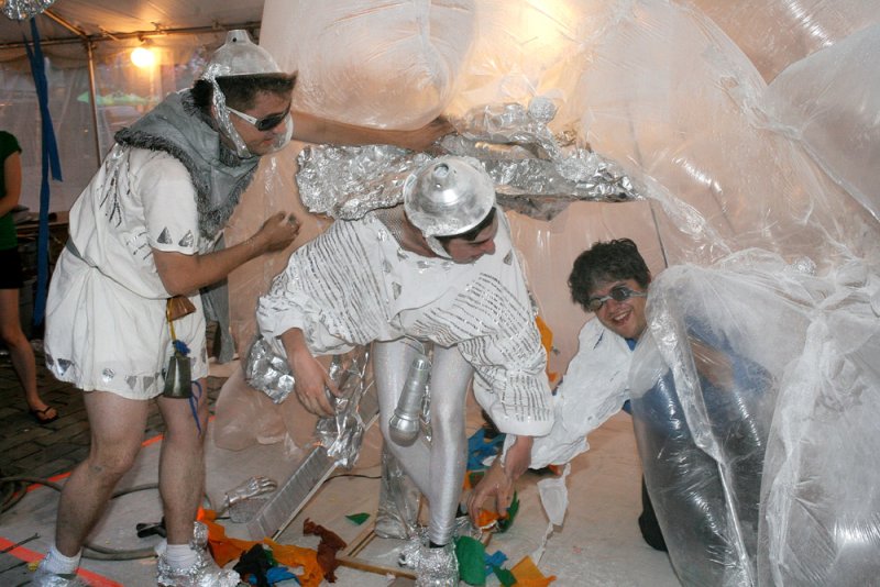 Three performers in silver and white costumes interact with a large, clear inflatable structure, surrounded by colorful debri