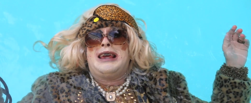 Blonde person in drag, wearing a leopard print coat, headband, and sunglasses, floating in bright blue water.