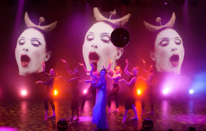 Performance with dancers on stage, illuminated by pink and orange lights, in front of three large projected faces with open mouths.