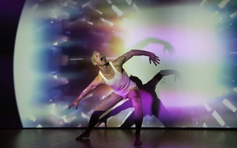 A male dancer in a white tank top and briefs, mouth open, strikes an expressive pose against a vibrant, projected backdrop of swirling colors.