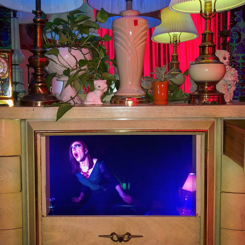 A woman with dark hair and red lipstick screams, illuminated by blue light, in a framed video display below lamps and plants.