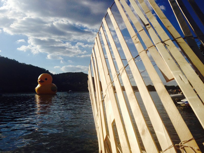 Giant yellow rubber duck floating on water, with a wooden slatted structure in the foreground under a cloudy blue sky.