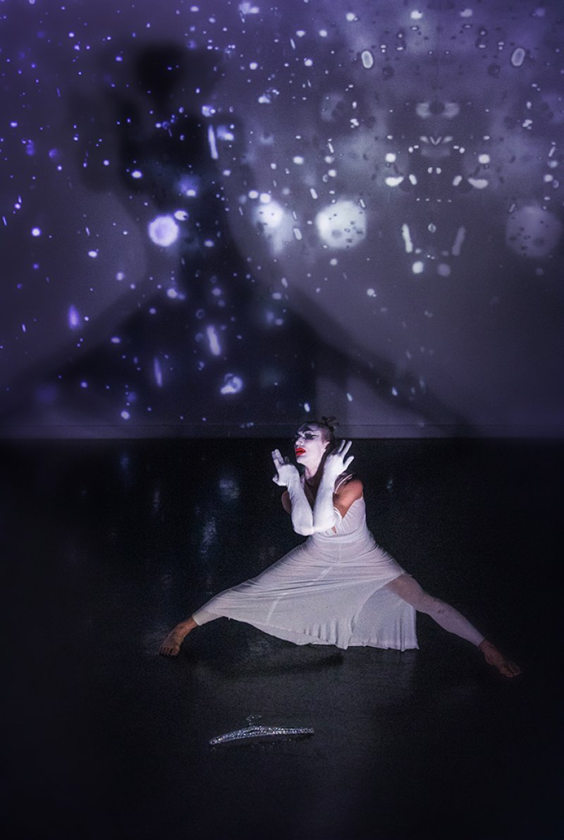 Performer in white dress and gloves, with clown makeup, poses on dark floor under purple projected light with white spots.