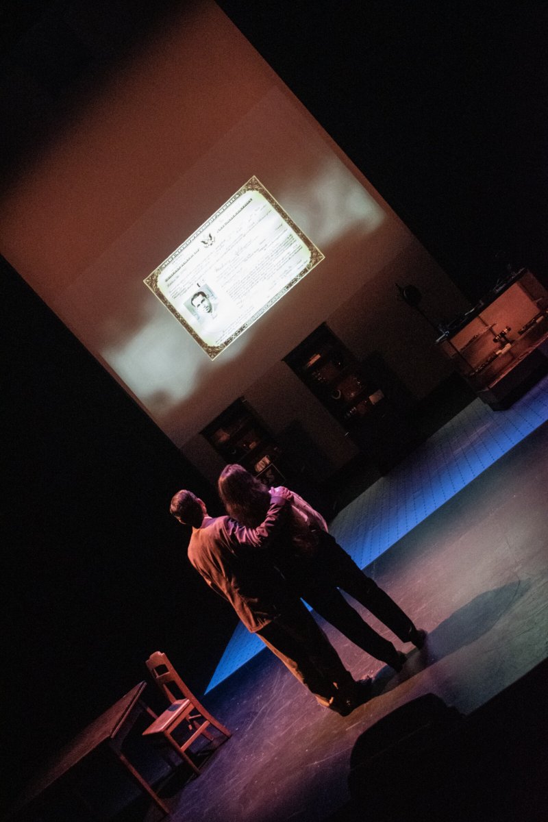 Two figures on a stage gaze at a projected document, illuminated by a blue light.
