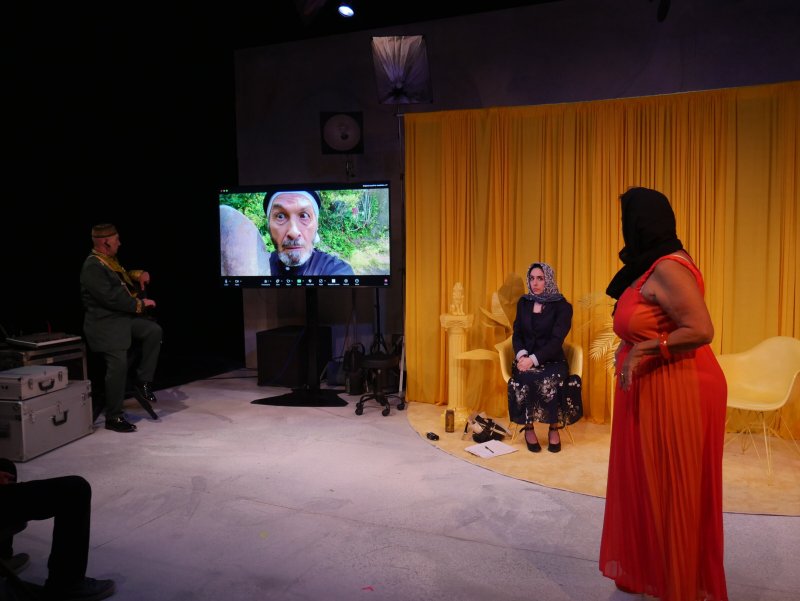 Stage scene with three actors: one on a screen, one seated, and one standing in an orange dress.