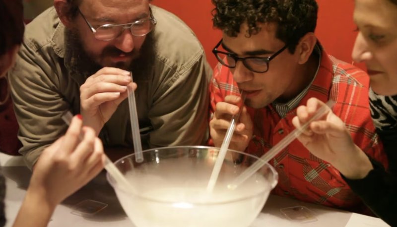 Four people with straws blowing into a bowl of dry ice vapor, red background.
