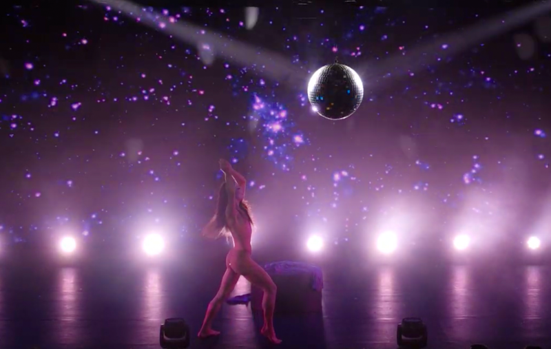A nude figure with long hair dances under a disco ball, illuminated by purple stage lights and a starry background.
