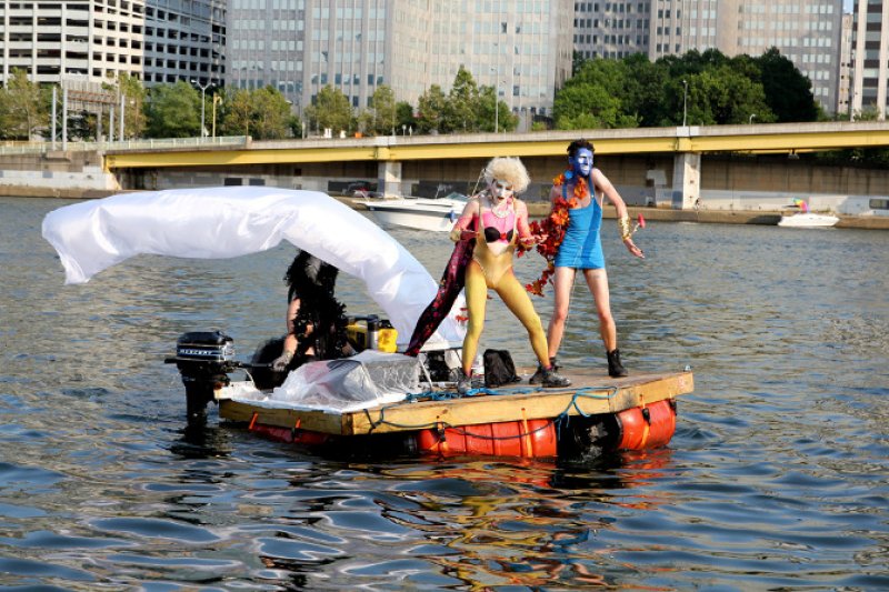 Performance art with two costumed figures on a small raft with an outboard motor, a white parachute, and city buildings in th