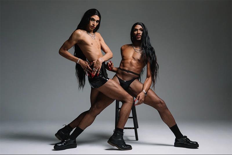 Two drag queens with long black hair, one in a black vinyl skirt and fishnets, the other in black briefs and fishnets, posing against a gray background.