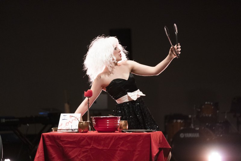 Performer in white wig, black sequin dress, and white bow, holding tongs, looking at reflection in tongs. Red table, dark bac
