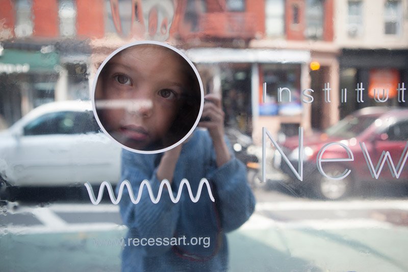 Child's face in a circular reflection on a glass window, with blurred city street and buildings in the background.