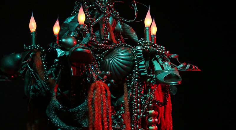Dark sculpture of shoes, beads, and objects with four glowing orange candle-like lights against a black background.