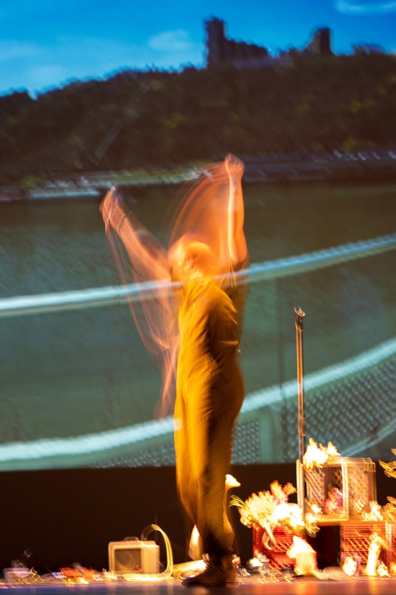 Blurred figure in yellow-orange light with arms raised, performing in front of a projected landscape with a castle.