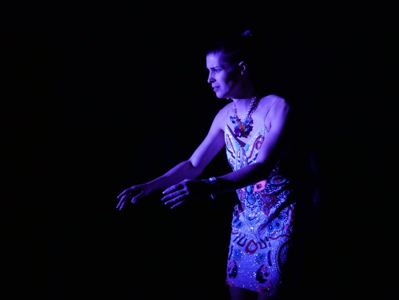 Woman in a colorful, beaded dress and statement necklace, illuminated by purple light against a dark background, extending he