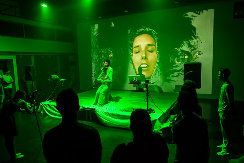 Live performance with a man on stage under green light, a large projected face behind him, and silhouetted audience.
