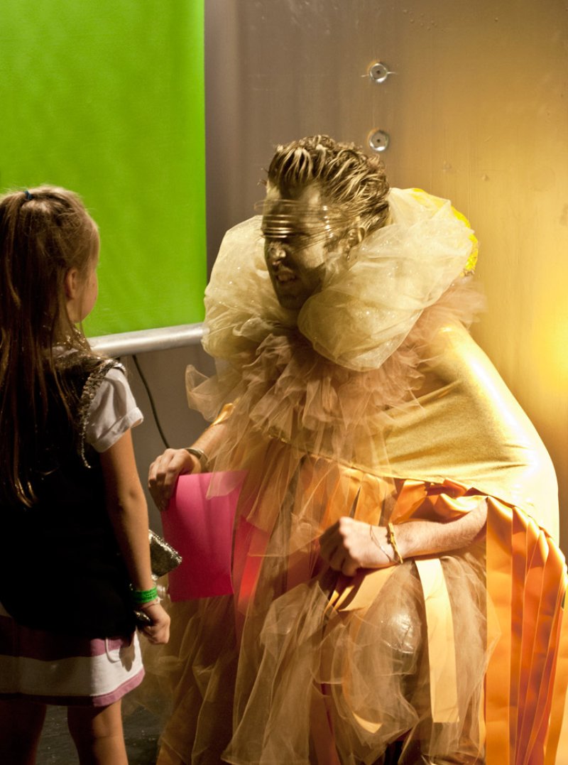 Performance artist in gold body paint and elaborate ruffled costume interacts with a young girl, against a bright green and y