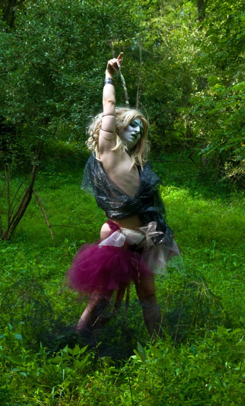 Performer in a dark mesh top, magenta tutu, and white face paint, with an arm raised, in a lush green forest.