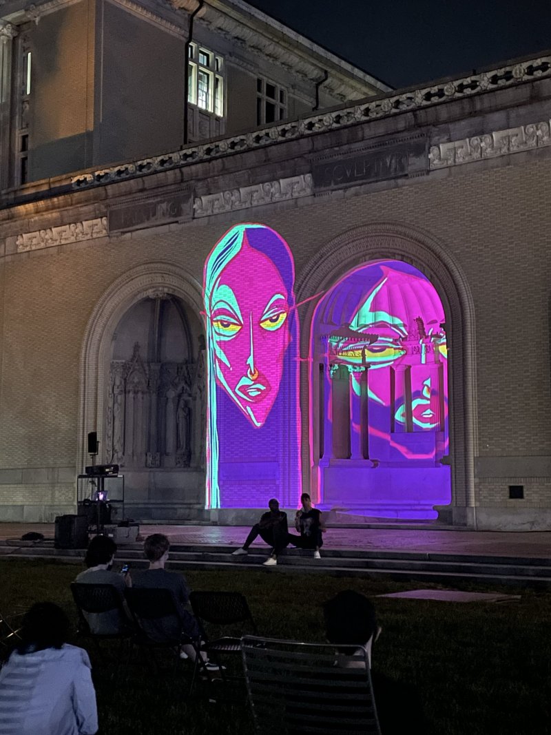 Two large, neon-colored digital portraits projected onto a brick building at night, with people sitting on a lawn watching.