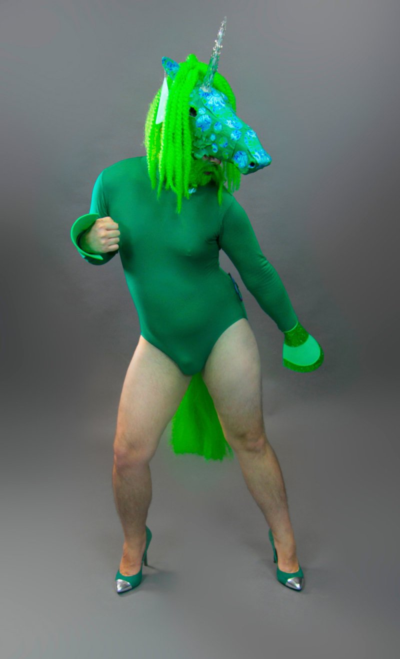 Person in green bodysuit and heels, wearing a blue unicorn mask with bright green hair and tail, poses against a gray backgro