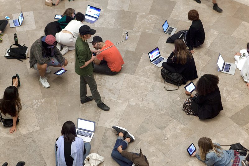 Overhead view of people sitting on a tiled floor, engrossed in laptops, tablets, and phones.