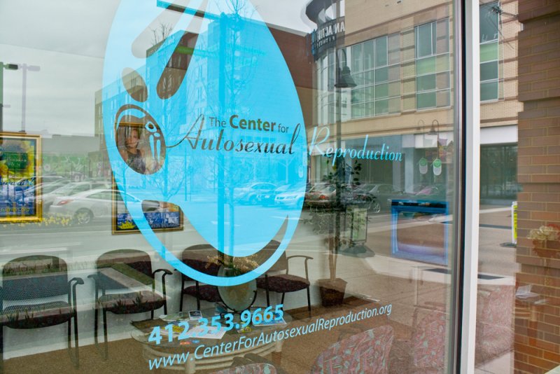 Light blue oval sticker on a glass door, reflecting a street and building, with text "The Center for Autosexual Reproduction"