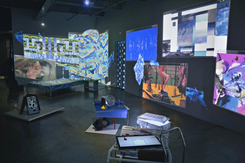 Multimedia installation with screens displaying abstract blue, purple, and white digital art, and various objects on the floo