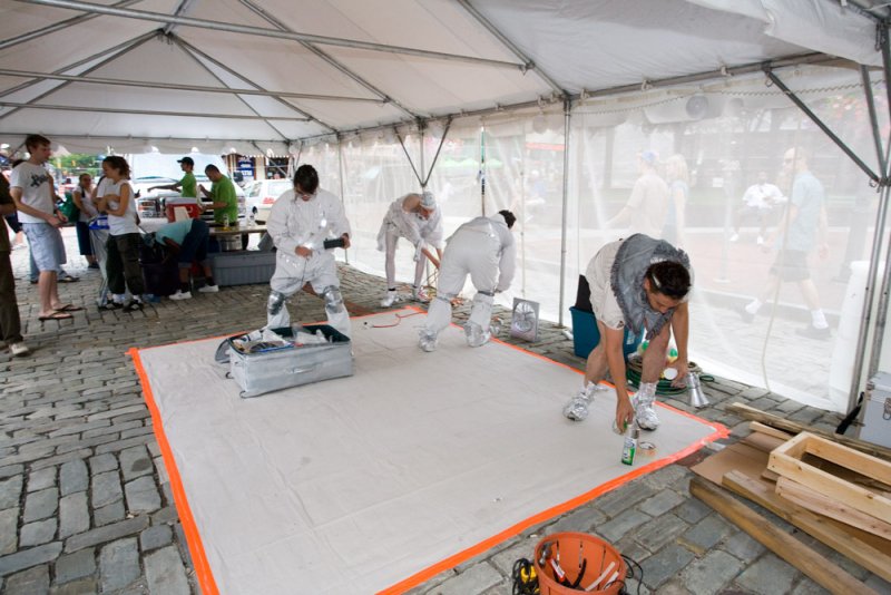 Performance art under a white tent with people in white hazmat suits and silver boots working on a white floor mat.