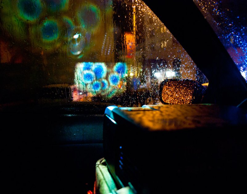Night scene through rain-streaked car window; blurry lights, side mirror, dark interior.