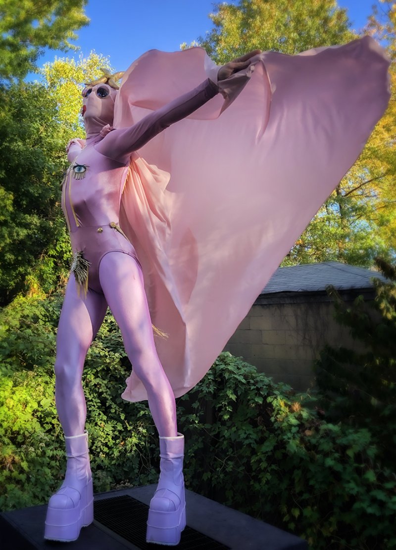 A person in a full pink bodysuit, mask, and platform boots with a large pink cape outstretched against a vibrant green and ye