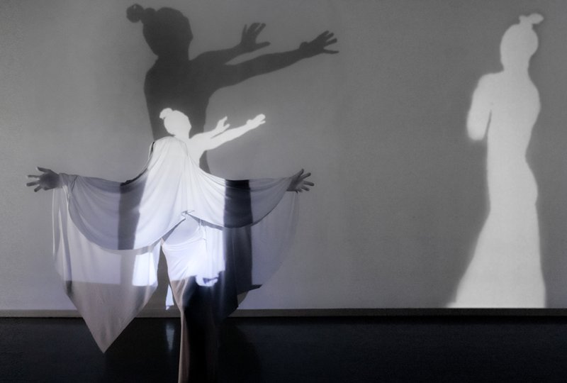 A figure in white flowing fabric with outstretched arms, casting a large dark shadow and a smaller white shadow on a gray wal