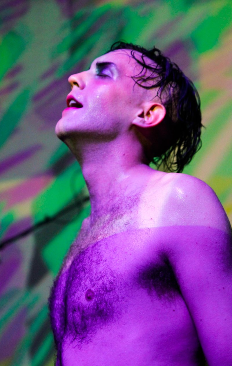 Purple-lit, bare-chested person with wet hair, looking up. Green and pink background.