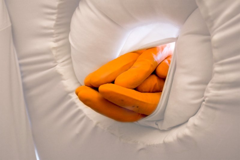 Orange-gloved hands emerge from a white, padded, circular opening, suggesting a sterile or contained environment.