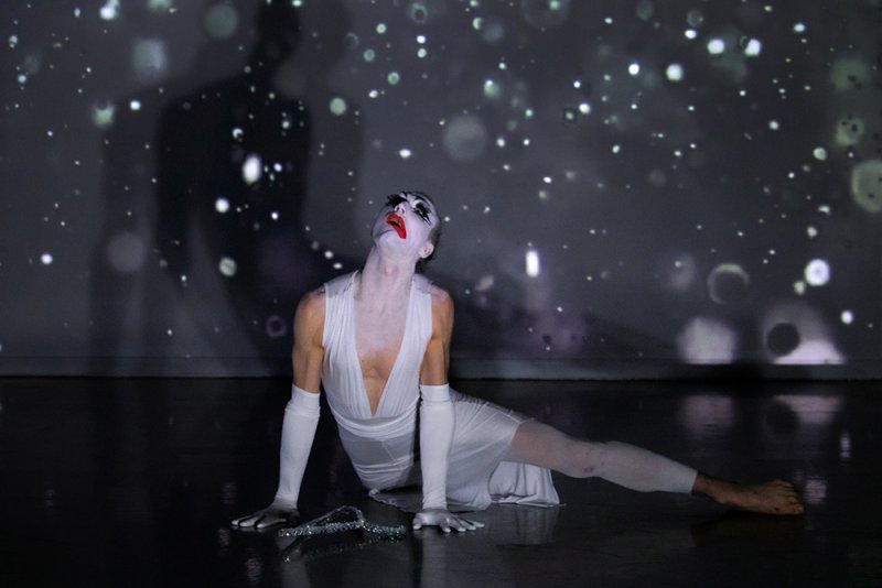 Performer in white with clown makeup, red lips, and glitter, looking up against a dark background with projected white lights
