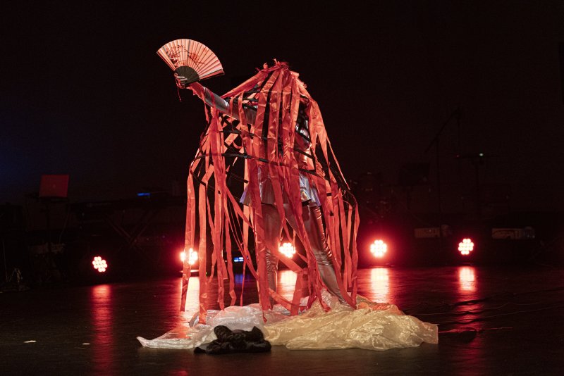 Performance art: Figure in silver suit with red streamers, holding fan, on dark stage with lights.