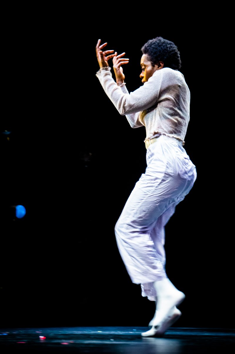 A dancer with dark skin and curly hair, wearing a shimmering top and light pants, leaps against a black background.