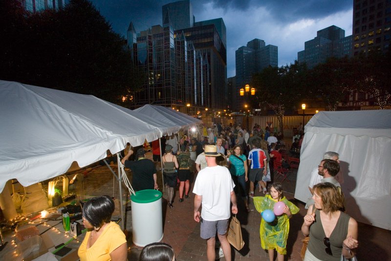 Nighttime outdoor event with white tents, crowds of people, and city buildings under a dark, cloudy sky.