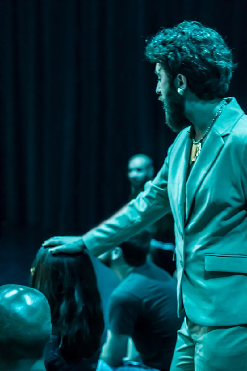 Man with dark curly hair and beard, wearing a light green suit, hand resting on another person's dark hair.