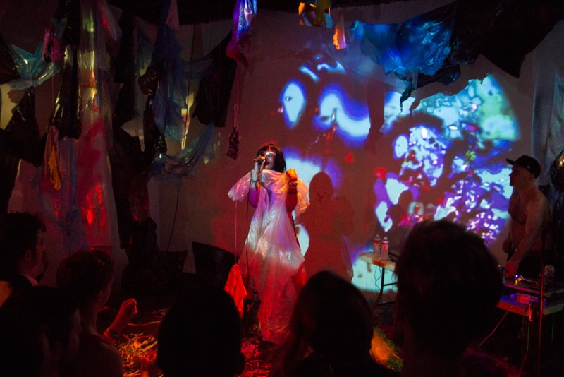 Performance art with a person in a white dress singing into a mic, illuminated by blue and purple projections.