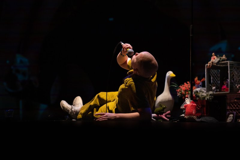 Bald performer in yellow jumpsuit reclines on dark stage, singing into a microphone, with a white duck prop behind them.