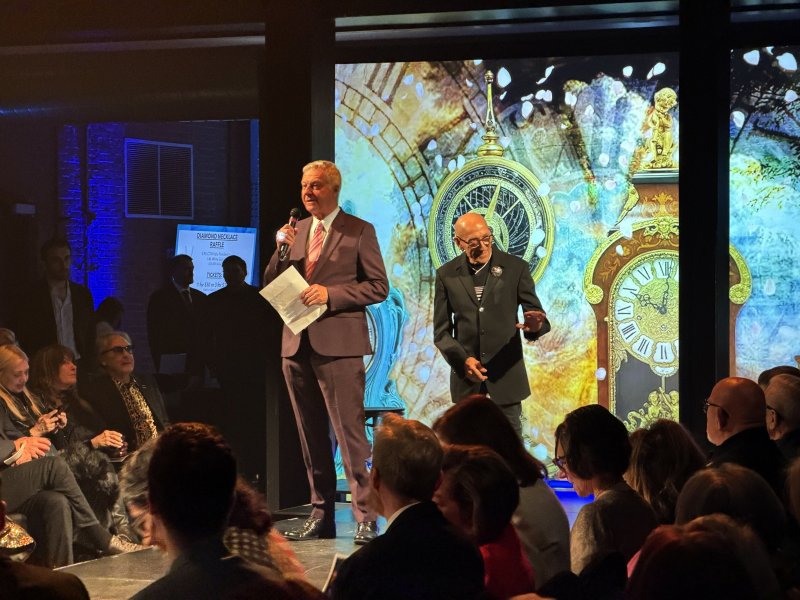 Two men on stage, one speaking into a microphone, in front of a large screen displaying ornate clocks, with an audience watching.