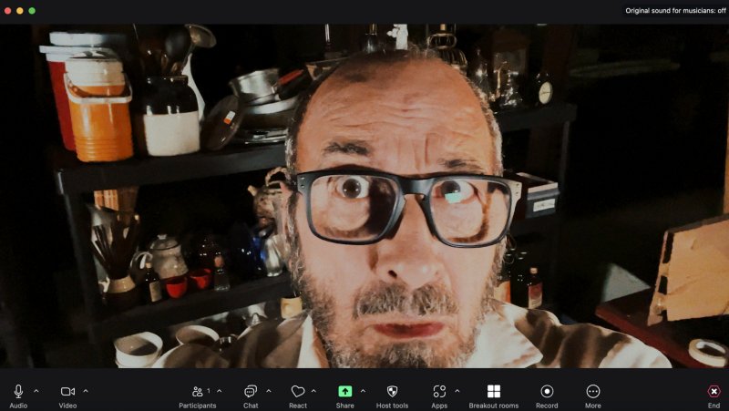 Man with wide eyes and glasses in a video call, surrounded by a cluttered shelf with various objects.