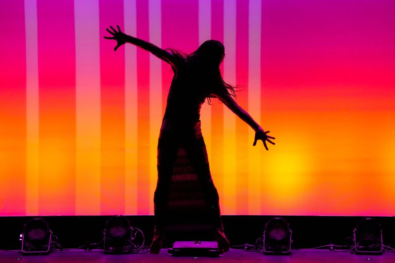 Silhouette of a dancer in a dress against a vibrant orange and pink striped background.