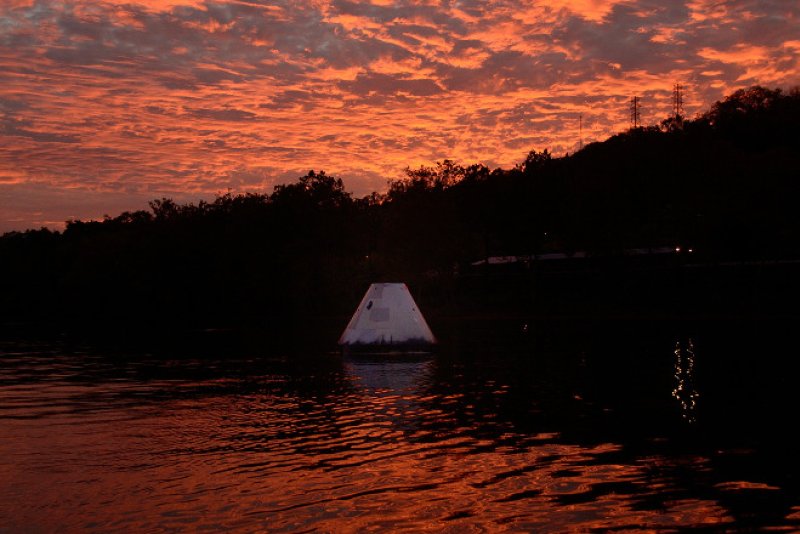 A white space capsule floats in dark water under a dramatic orange and red sunset sky, with a silhouetted treeline and hill i