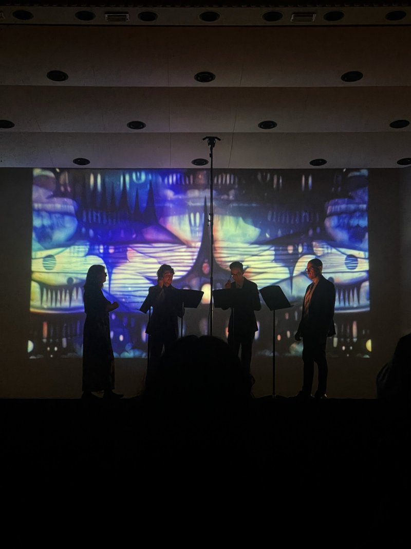 Four silhouetted figures on stage in front of a large screen displaying abstract, colorful digital art.
