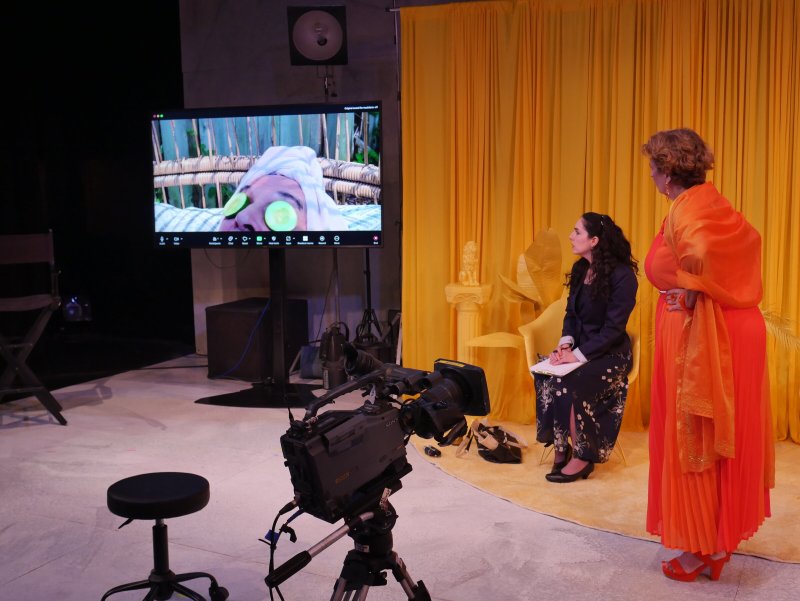 Two women on a set with yellow curtains, a large screen showing a person with a face mask, and a camera in the foreground.