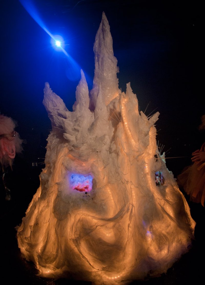 Sculptural installation resembling a jagged, glowing white mountain with orange and blue internal lights against a dark backg