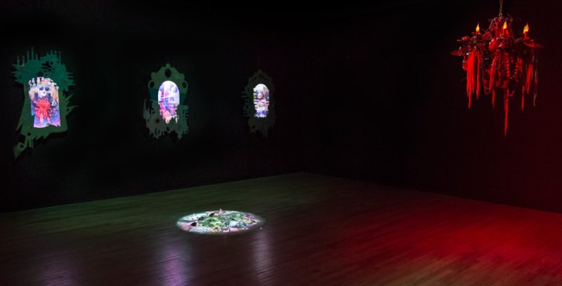 Dark room with three glowing, ornate mirror-like screens on a black wall, a circular projection on the floor, and a red chand